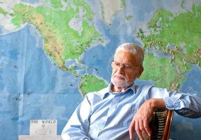 Man with white hair sits in front of a world map Man with white hair sits in front of a world map